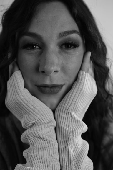 Portrait einer Person mit langen, lockigen Haaren, die nachdenklich mit den Händen das Gesicht stützt.
